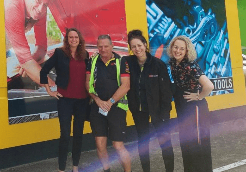 Graham is in from of a Repco store wall on the outside with Ashlee to the left, his manager Erin to the right and another Help team member next to Erin. They are all looking and smiling at the camera.