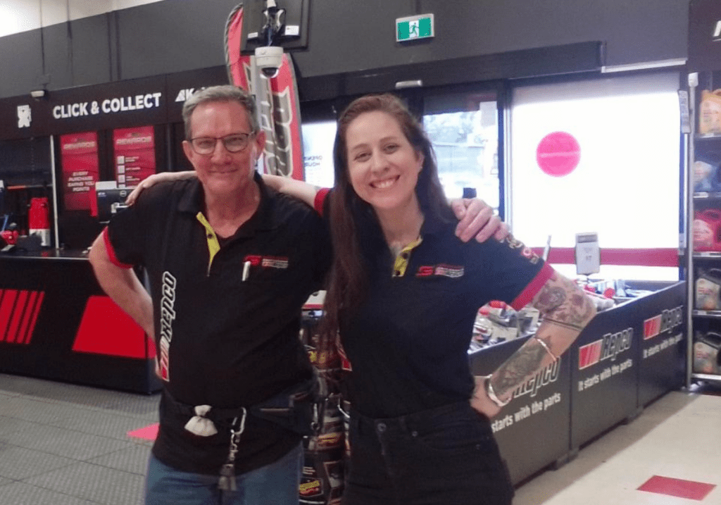 Graham with his left arm around a colleague's shoulders while both smile at the camera in Repco store.