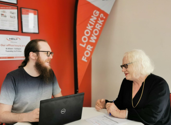 Sam engaged in a discussion with a client during an employment coaching session, both sitting at a table with notebooks and resources, focusing on career development.